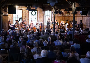 Blick auf eine große Musikgruppe in Tracht auf der Bühne vor vollem Saal. | © Hans Fuchs