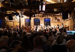 Blick auf eine große Musikgruppe in Tracht auf der Bühne vor vollem Saal. | © Hans Fuchs