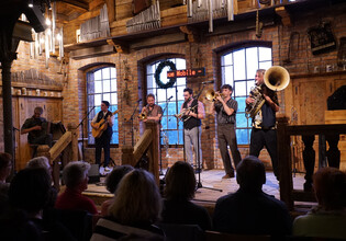 Musikband mit Blasinstrumenten und Gitarren auf einer Bühne vor großen Fenstern. | © Hans Fuchs