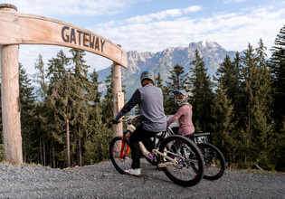 Zwei Mountainbiker:innen mit Fullface-Helmen stehen mit ihren Bikes unter dem hölzernen „Gateway“-Bogen am Start eines Trails im Bikepark Leogang mit Blick auf das Steinerne Meer. | © Yvonne Hörl