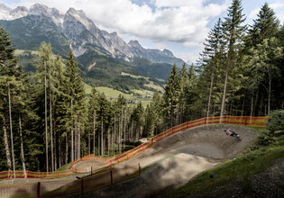 Mountainbiker in einem großen Anlieger der T-Line, die sich durch den Wald schlängelt. Im Hintergrund das beeindruckende Panorama der Leoganger Steinberge. | © Yvonne Hörl