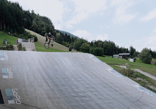 Ein Biker schiebt sein Rad über den Schotterweg in Richtung Bagjump-Rampe im Übungsareal bei der Talstation Leogang. Rechts ist das große GoPro-Luftkissen für sichere Landungen zu sehen. | © Yvonne Hörl